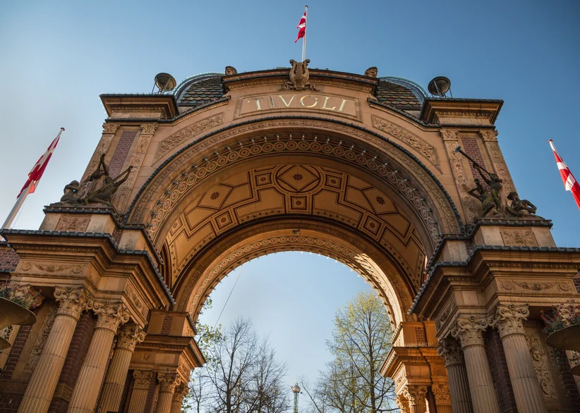 Low-angle view of Tivoli Gardens' ornate stone entrance arch bearing the name 'Tivoli,' flanked by decorative carved figures and Danish flags, with bare trees visible through the archway against a clear blue sky.
