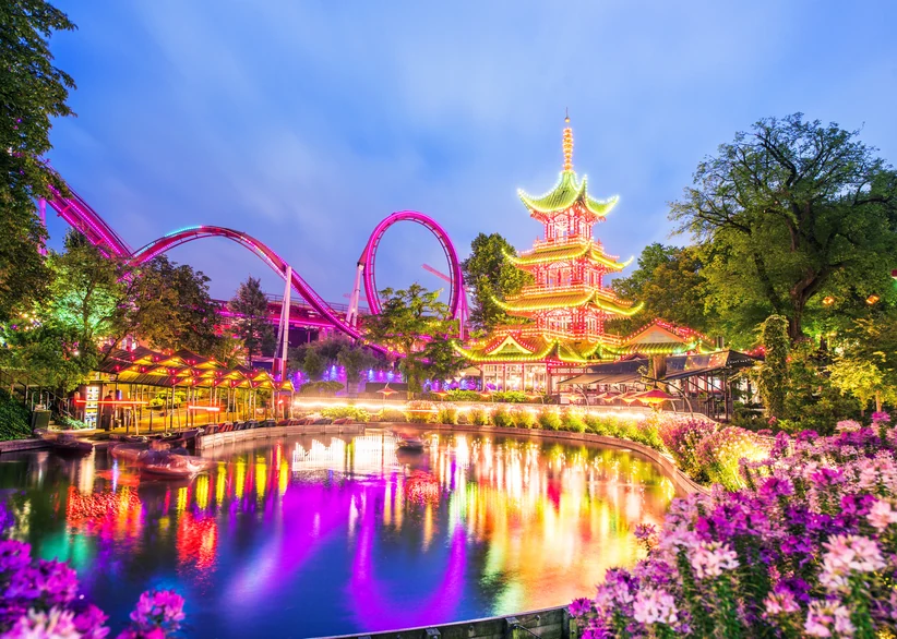 Illuminated Tivoli Gardens at dusk, with a pink roller coaster looping beside a glowing multi-tiered Chinese-style pagoda, colorful lights reflecting on the lake, and pink flowers blooming in the foreground.
