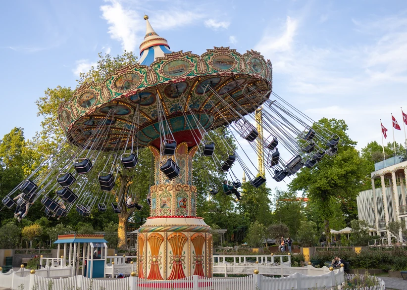 A traditional swing carousel ride spinning riders in chair-swings beneath a decorative canopy at Tivoli Gardens, surrounded by lush trees, with Danish flags and a white pavilion visible in the background under a partly cloudy sky.