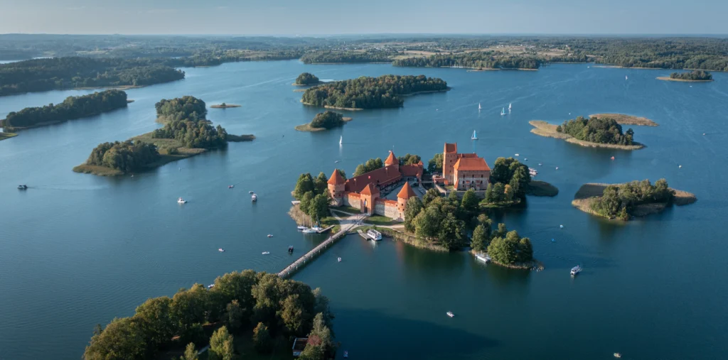 Aerial view of Trakai Island Castle's red-brick towers on a small forested island connected by a wooden footbridge, surrounded by a blue lake dotted with sailboats and small green islands.