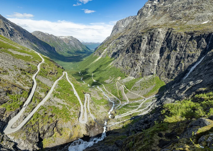 Aerial view of Trollstigen's dramatic hairpin switchback road winding down a steep green valley between towering rocky mountains, with a waterfall and stream running alongside the road.