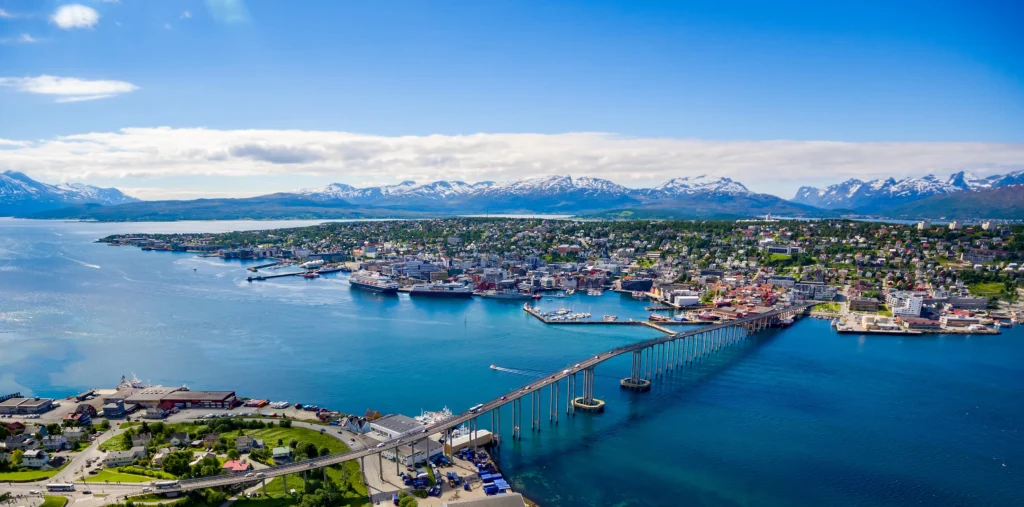 Aerial view of the Tromsø Bridge spanning the strait between the city center and Tromsdalen, with harbor cruise ships docked along the waterfront and snow-capped mountains rising in the distance