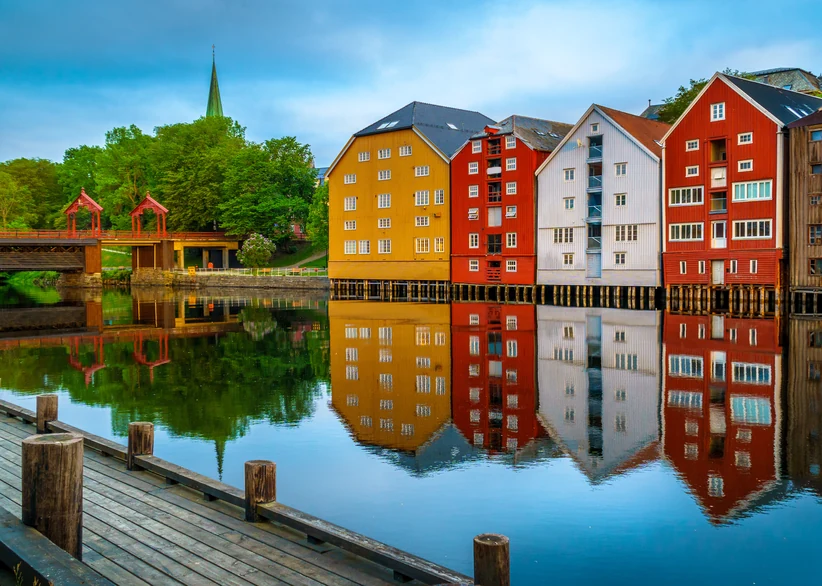 Colorful old wooden warehouse buildings in yellow, red, and white line the Nidelva river in Trondheim, reflecting on the calm water beside a wooden pier, with a church spire and red footbridge visible in the background.