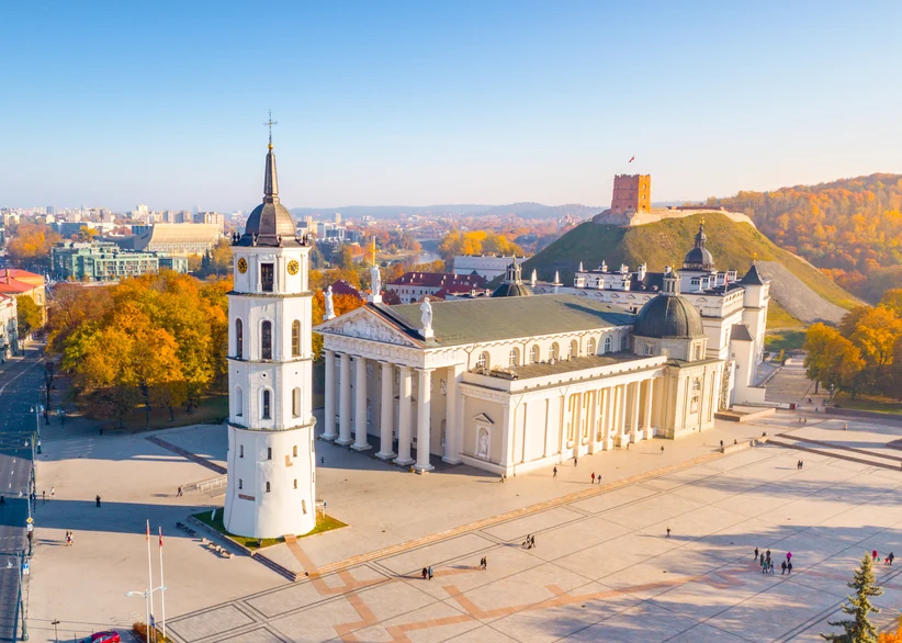 Vilnius Cathedral's white neoclassical facade and freestanding bell tower stand in Cathedral Square, with Gediminas' Tower visible on a forested hill in the background under a clear autumn sky.