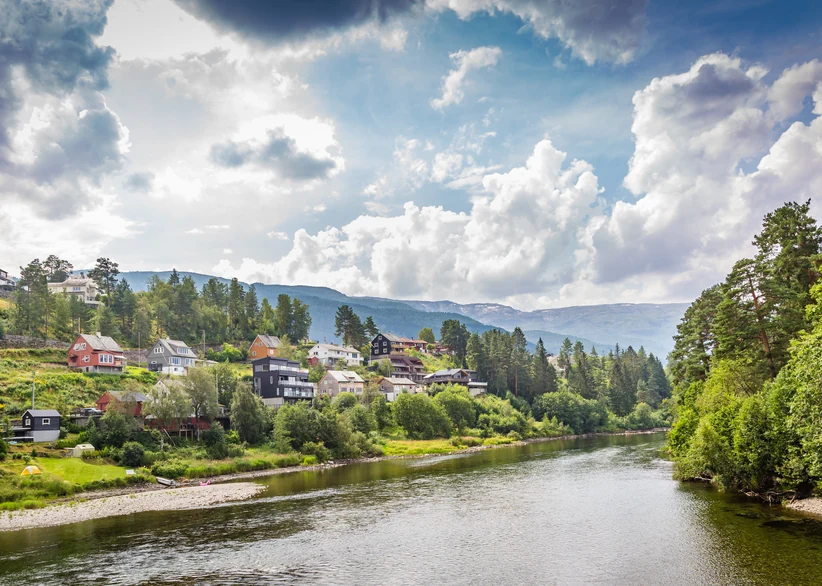 Colorful houses of the town of Voss dot a green forested hillside overlooking a calm river, beneath a dramatic sky filled with billowing clouds and distant mountains.