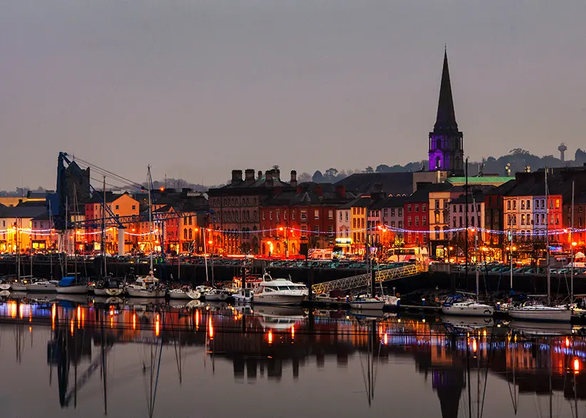Waterford's colourful quayside buildings and marina lit up at dusk with festive lights, a church spire, and boats reflected in the still River Suir, Ireland