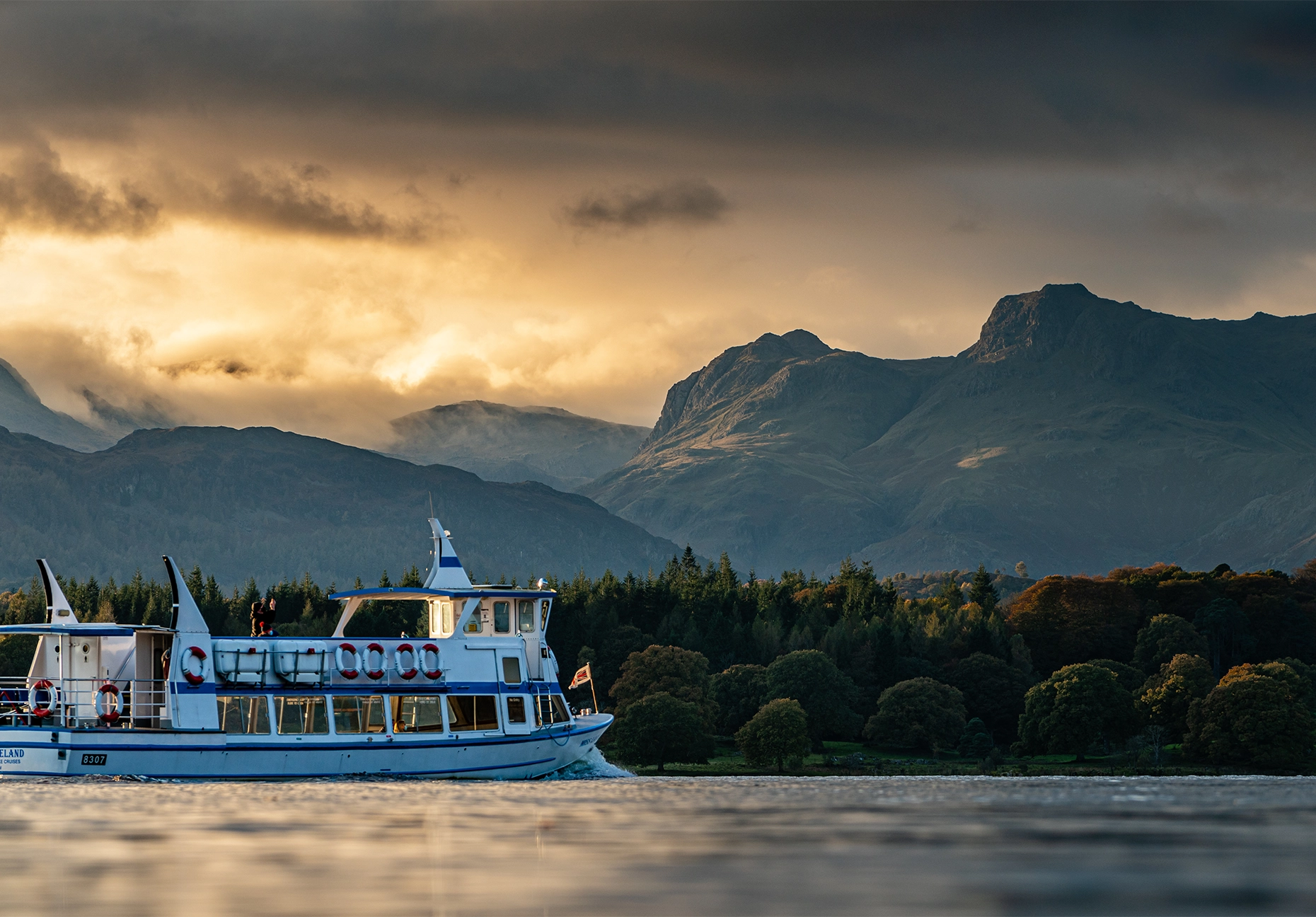 A cruise boat crosses Windermere at sunset, with the Langdale Pikes rising behind the treeline.