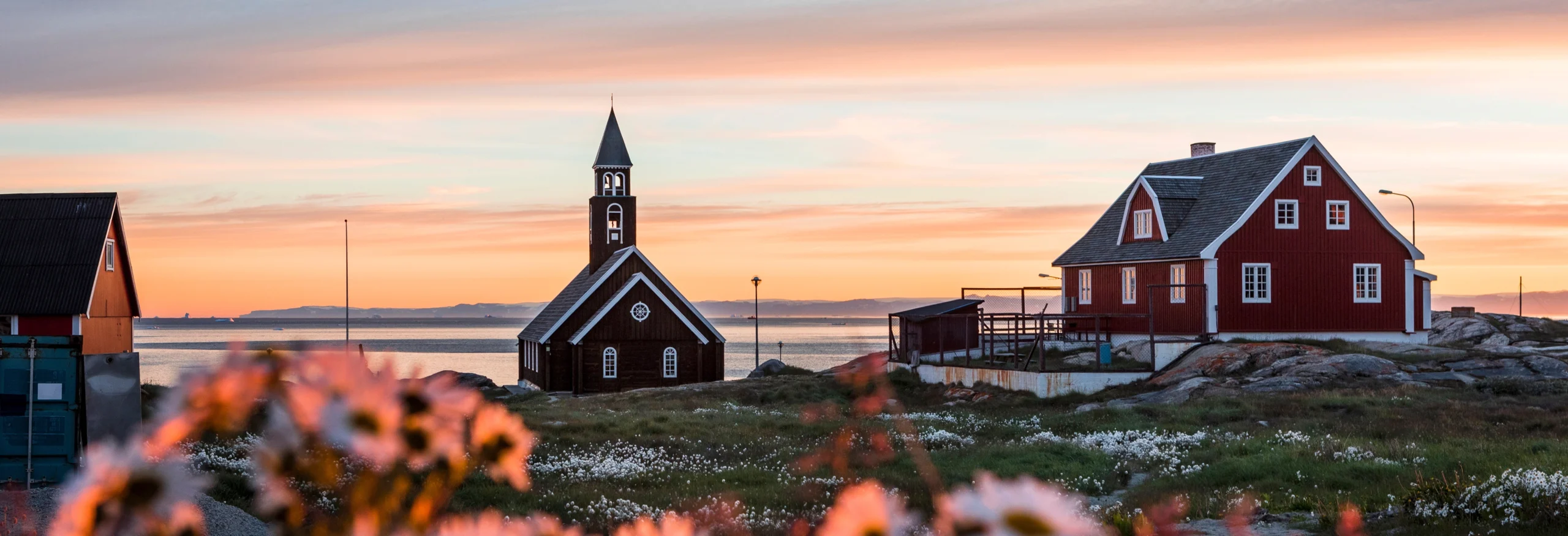 Panoramic sunset view of Ilulissat's wooden church, red house, and coastal wildflower meadow.