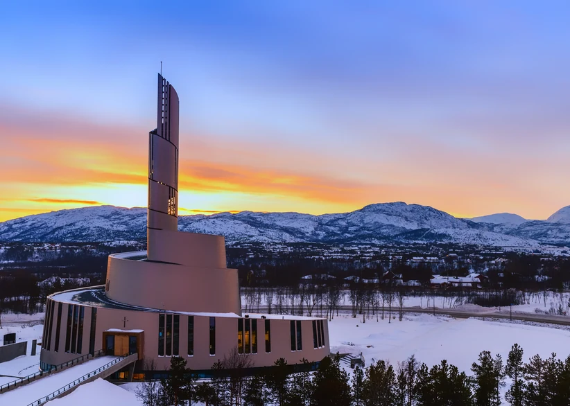 The striking modern spiral spire of Alta's Northern Lights Cathedral stands against a vivid orange and blue sunset sky, with snow-covered mountains and a frozen landscape in the background.