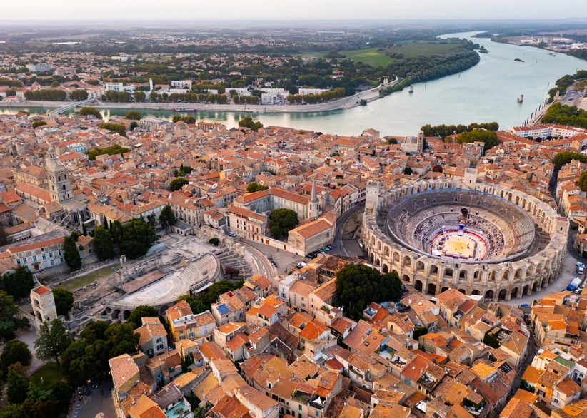 Aerial view of Arles' Roman amphitheatre and ancient theater ruins amid the town's orange-tiled rooftops, with the Rhône river curving through green farmland in the background at dusk.