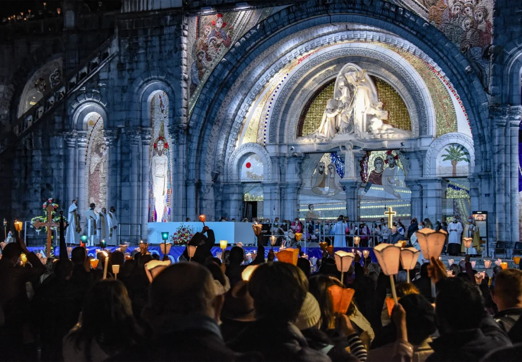 Pilgrims raise lit candles before the mosaic-covered Rosary Basilica facade during an evening Mass.
