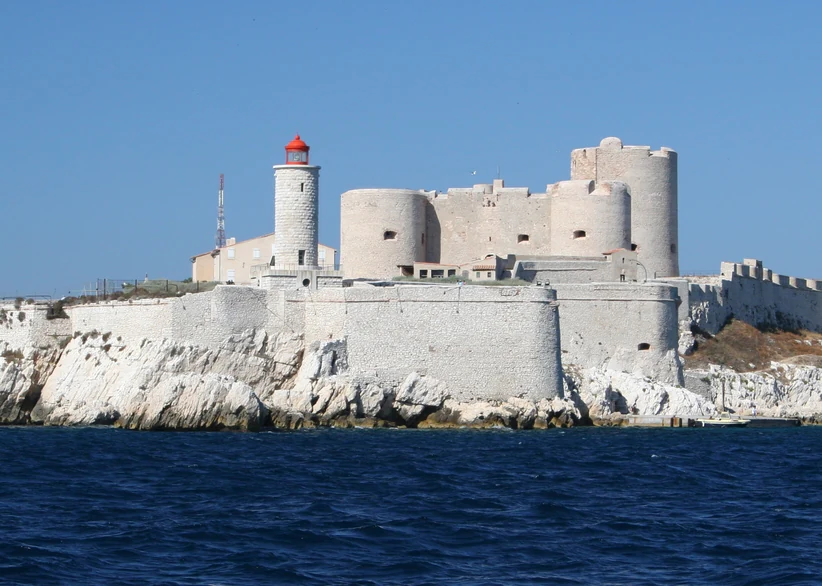 Château d'If's round white stone towers and red-capped lighthouse sit atop a rocky island fortress, surrounded by fortified walls and deep blue sea under a clear sky.