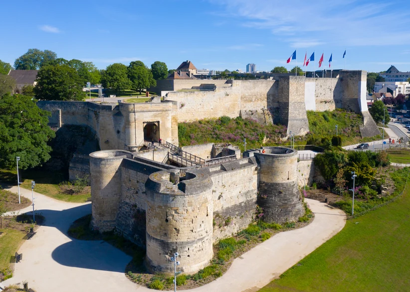 Aerial view of the medieval Château de Caen's stone ramparts and round towers, with French and regional flags flying atop the walls and the modern city skyline visible in the background under a clear blue sky.