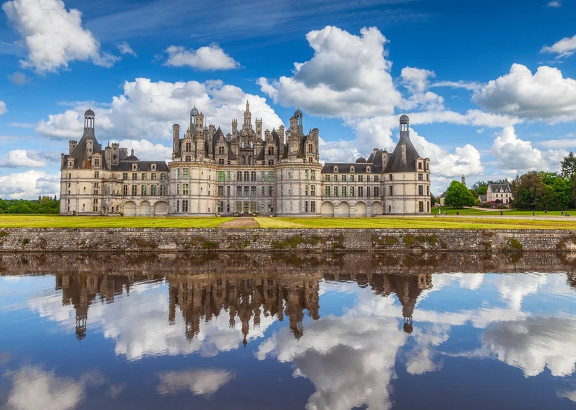 Château de Chambord's elaborate Renaissance towers, turrets, and chimneys reflect perfectly in the still moat water, framed by manicured lawns and a dramatic sky of billowing white clouds.