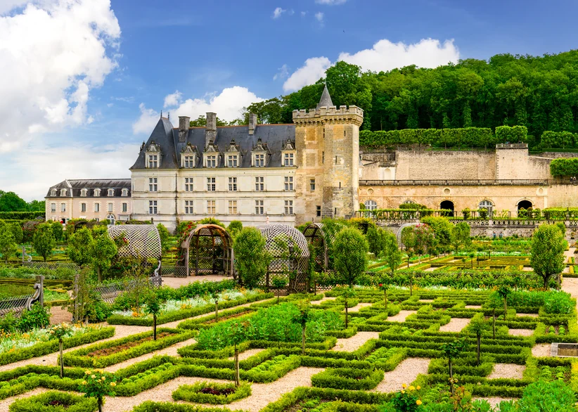 The formal boxwood-hedged gardens of Château de Villandry stretch before the turreted château, with trellised arbors and neat rows of vegetables and flowers under a partly cloudy sky.
