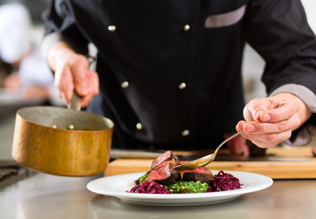 A chef in black whites pours sauce from a copper pan over sliced steak with red cabbage.