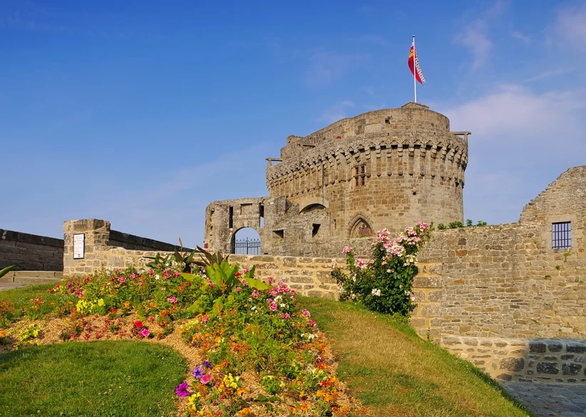 The round stone keep of a medieval castle flies a red and white flag above weathered fortress walls, with colorful flowerbeds and climbing roses lining the grassy courtyard under a clear blue sky.