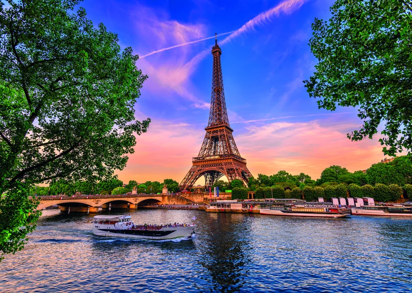 The Eiffel Tower rises above the Seine river at sunset, framed by green trees on either side, with a tour boat cruising past and a bridge crossing the water beneath a vivid pink and orange sky.