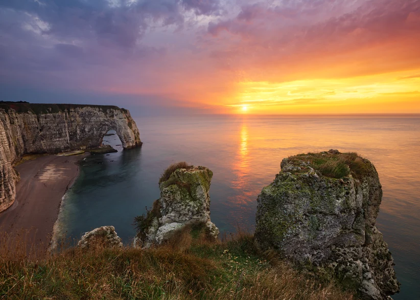 Sunset over Étretat's white chalk cliffs and natural rock arch, with lichen-covered rock formations in the foreground and the sun's golden glow reflecting across the calm sea beneath a dramatic pink and orange sky.