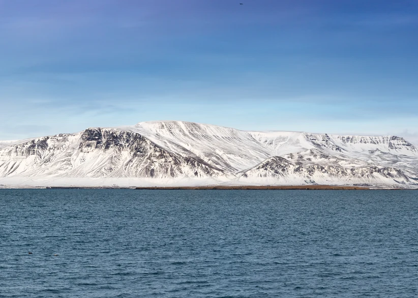 A snow-covered mountain ridge with deep vertical striations rises above the calm blue waters of Faxaflói Bay under a clear sky.