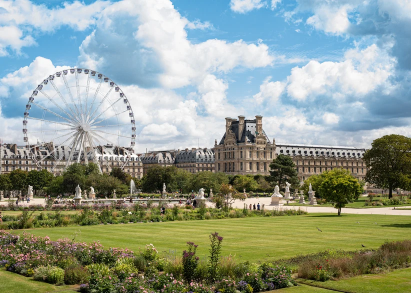 A Ferris wheel and the Louvre's ornate roofline rise beyond the manicured lawns, flowerbeds, and classical statues of the Jardin des Tuileries under a partly cloudy blue sky.