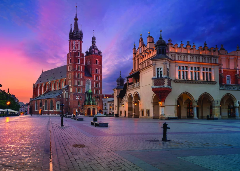 St. Mary's Basilica with its two asymmetrical towers and the Renaissance Cloth Hall glow at dawn on Kraków's Main Market Square, beneath a vivid pink and purple sky with wet cobblestones reflecting the light.