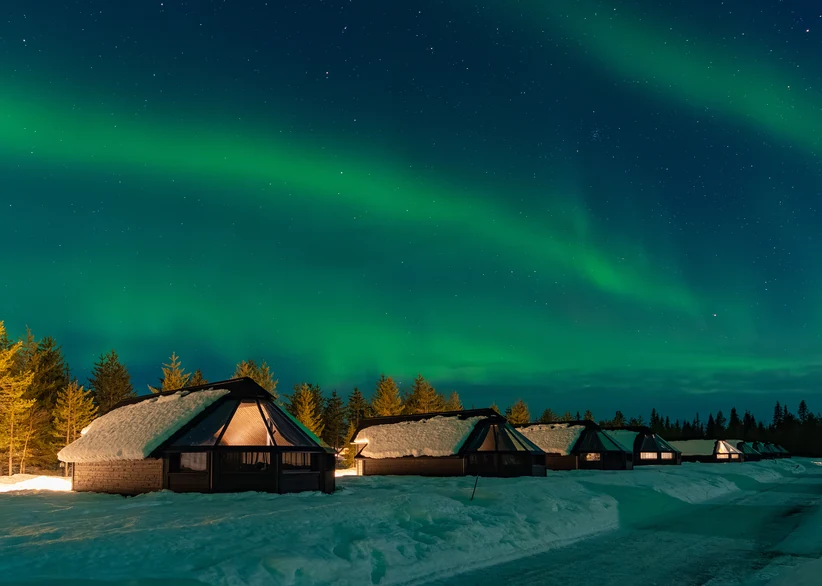 Vivid green aurora borealis ribbons swirl across a starry night sky above a row of snow-topped glass-roofed igloo cabins bordered by pine trees.
