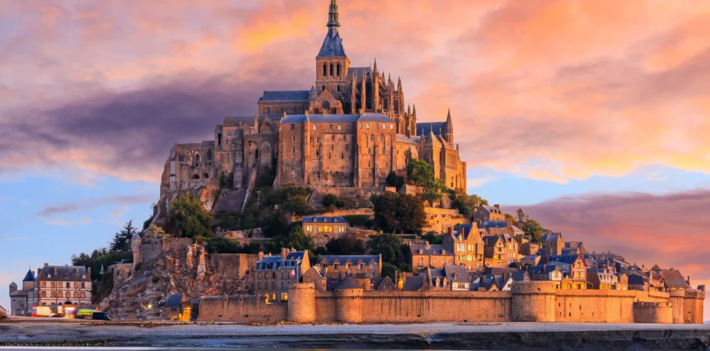 Mont Saint-Michel's spired abbey and fortified walls glow at sunset above the surrounding town and tidal flats, reflected in the still water beneath a dramatic sky of pink and purple clouds.