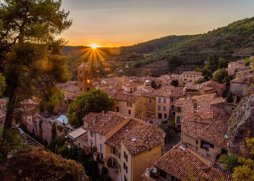 Sunset over the hilltop village of Moustiers-Sainte-Marie, with a stone bell tower and terracotta rooftops nestled among green hills, sunbeams bursting over the ridge in the distance.