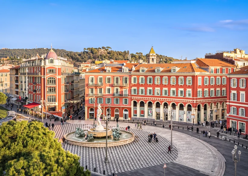 Aerial view of Nice's red-ochre Place Masséna, with its checkered pavement, central fountain statue, and arcaded buildings, backed by green hills under a clear blue sky.
