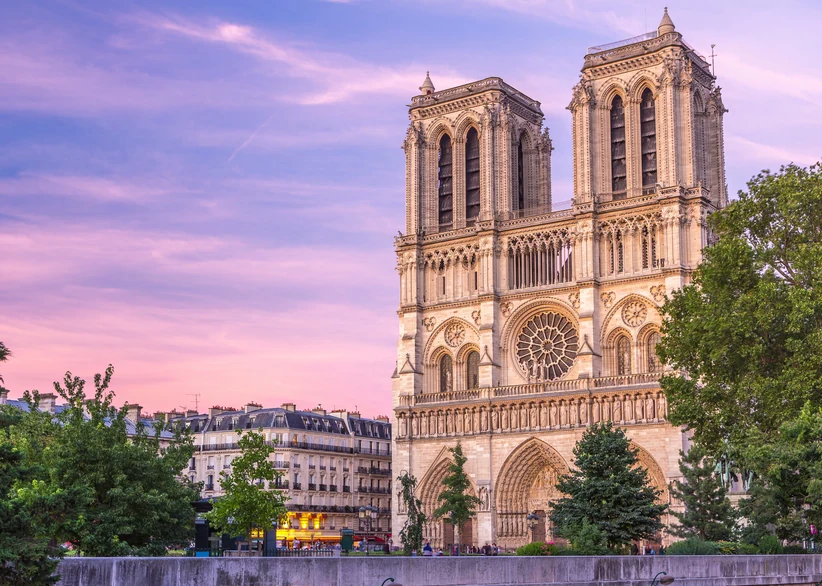 Notre-Dame Cathedral's twin Gothic towers and rose window rise above the trees at sunset, with the Parisian cityscape visible in the background beneath a soft pink and purple sky.