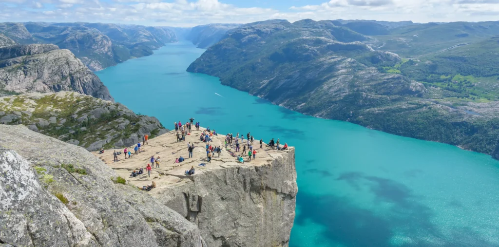 A wide aerial view of hikers gathered on the flat rock plateau of Preikestolen high above Lysefjord, its vivid turquoise water stretching between steep, forested mountains under a partly cloudy sky.
