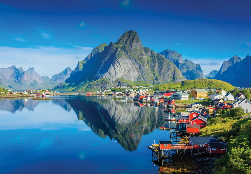 Red rorbu cabins line a calm fjord below jagged mountains reflected in still water.