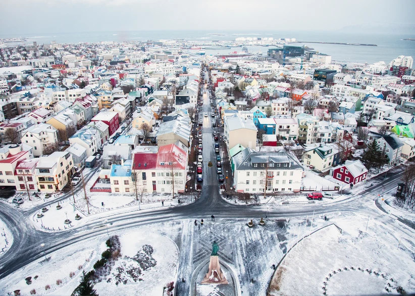 An aerial view down a snow-covered street in central Reykjavík lined with colorful rooftops, leading toward a statue and the harbor in the distance.