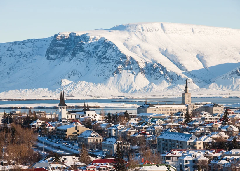 The spires of Hallgrímskirkja and Landakotskirkja rise above the snow-covered rooftops of Reykjavík, with the massive snow-capped Mount Esja looming across the bay.