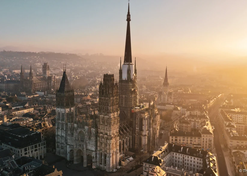 Aerial view of Rouen Cathedral's soaring Gothic spire and ornate facade rising above the city's rooftops at sunrise, with golden light and haze softening the skyline in the distance.