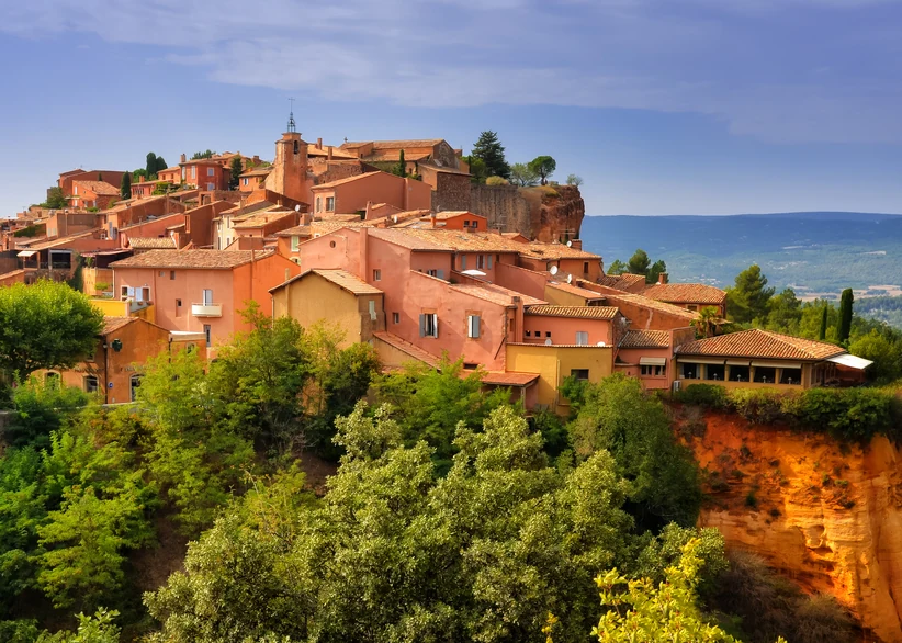 The ochre-colored village of Roussillon sits atop red cliffs, its terracotta rooftops and pastel orange buildings rising above lush greenery, with rolling hills stretching into the hazy distance.