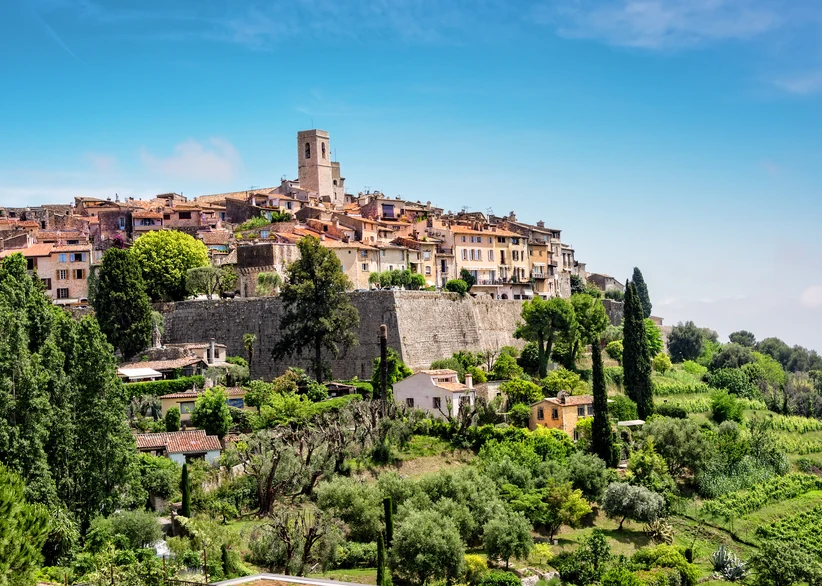 The fortified hilltop village of Saint-Paul-de-Vence rises above lush greenery and cypress trees, with its historic bell tower and ancient stone ramparts crowning the ridge under a clear blue sky