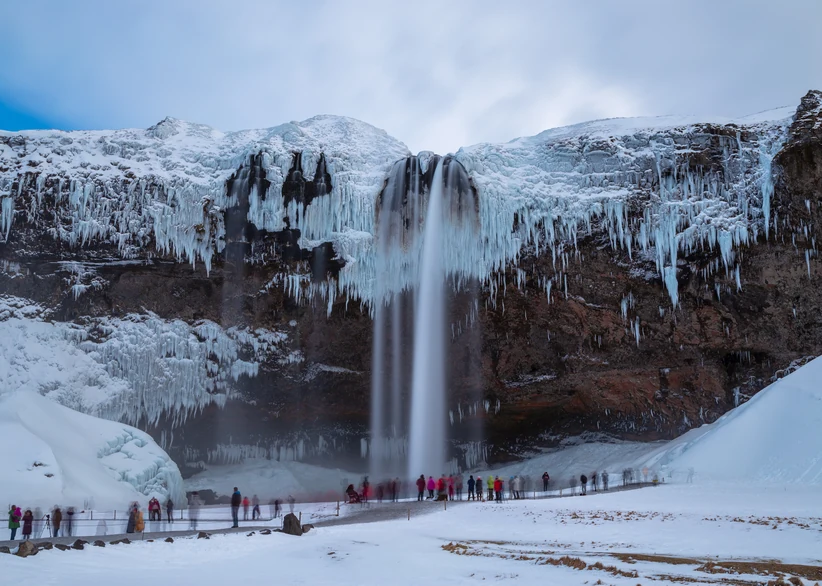 Seljalandsfoss waterfall cascades from an ice-draped cliff hung with massive icicles, as visitors in colorful winter clothing gather on the snowy path below.