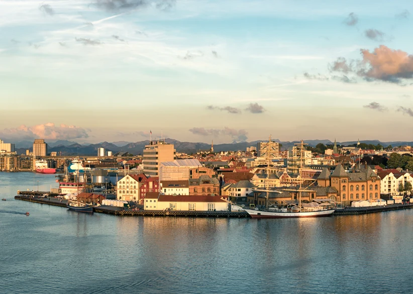 A waterfront view of Stavanger's harbor at golden hour, with colorful historic buildings, tall ships docked along the quay, and mountains visible in the hazy distance.