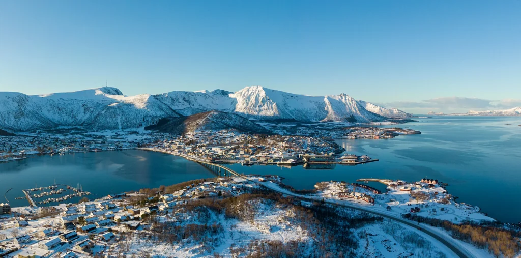 An aerial winter view of the town of Stokmarknes with its bridge crossing a calm blue fjord, surrounded by snow-covered houses and steep, snow-capped mountains under a clear sky.