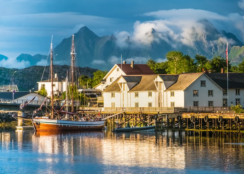 A wooden two-masted sailing ship moored at a pier in front of white multi-story waterfront buildings on pilings, with snow-dusted mountain peaks rising behind under a partly cloudy blue sky