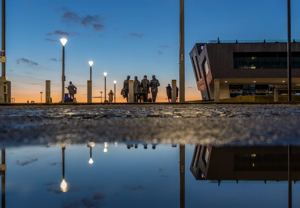 The Beatles statue silhouetted at dusk on Liverpool's Pier Head, mirrored in a rain puddle.