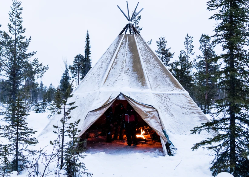 A snow-dusted traditional Sami tipi glows warmly from a campfire inside, nestled among snowy pine trees in the Lapland wilderness at dusk.