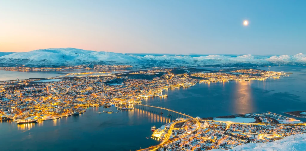 An aerial night view of Tromsø's illuminated city lights reflecting on the fjord, with the bridge connecting the islands and snow-covered mountains under a twilight sky with a bright moon.