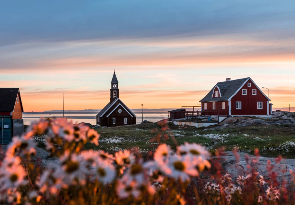 A wooden church and red timber house overlook the sea at sunset, framed by white daisies.