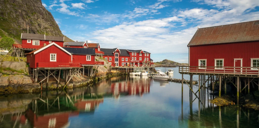 Traditional red wooden fishermen's cabins on stilts line the calm, clear waters of a harbor in the village of Å, with moored boats, reflections on the water, and a steep rocky mountainside under a blue sky with scattered clouds.