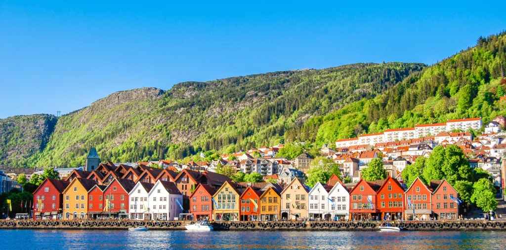 The colorful wooden gabled houses of Bergen's historic Bryggen waterfront stand along the harbor, with a forested green mountainside and hillside homes rising behind under a bright blue sky.
