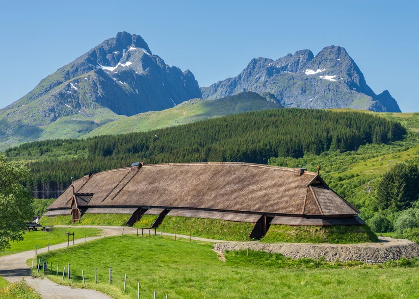 A reconstructed Viking longhouse with a turf-edged wooden shingle roof sits in a green meadow, with forested hills and rugged snow-streaked mountain peaks rising behind it under a clear blue sky.