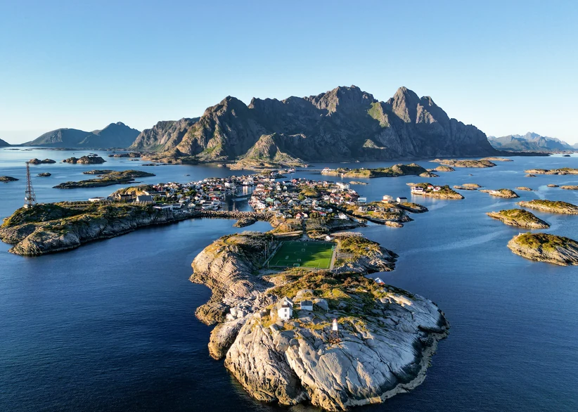 An aerial view of the fishing village of Henningsvær spread across small rocky islands, with its famous football pitch on a rocky outcrop in the foreground and dramatic jagged mountains rising behind calm blue waters.